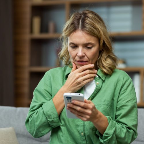 Close-up photo. Worried senior woman mother sitting on sofa at home and holding phone. Worries about children, writes and sends messages, calls, searches, waits at home.